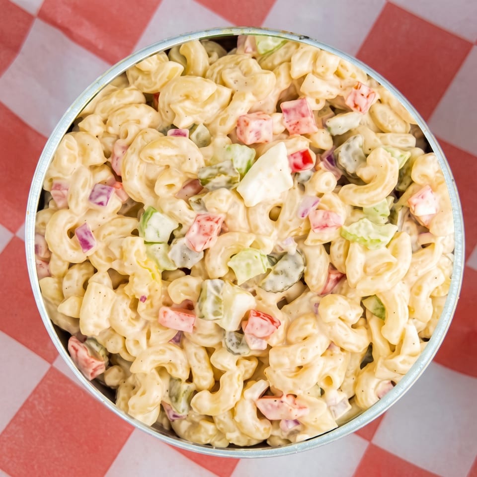 Creamy macaroni salad with red peppers, celery, and hard-boiled egg in a metal bowl.