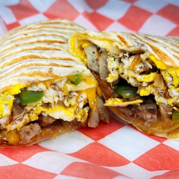 Grilled breakfast burrito, cut in half, revealing eggs, sausage, cheese, and green peppers on checkered paper.